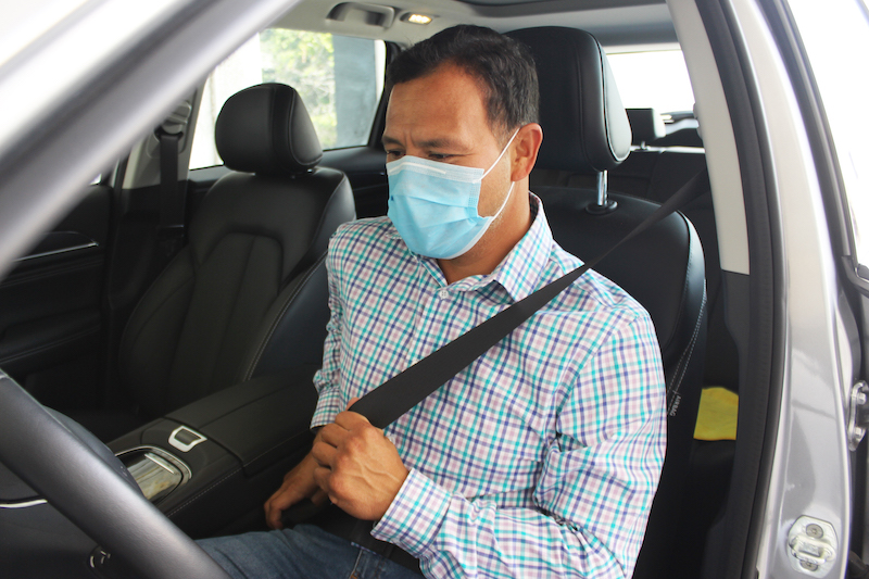 Conductor sentado dentro de un auto se coloca el cinturón de seguridad. Utiliza también un cubrebocas tricapa color celeste.